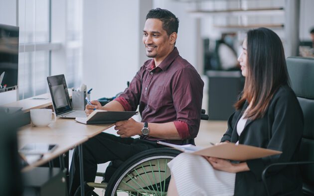 Two professionals sit at a desk in an office; one uses a wheelchair and writes in a notebook, while the other holds a clipboard.