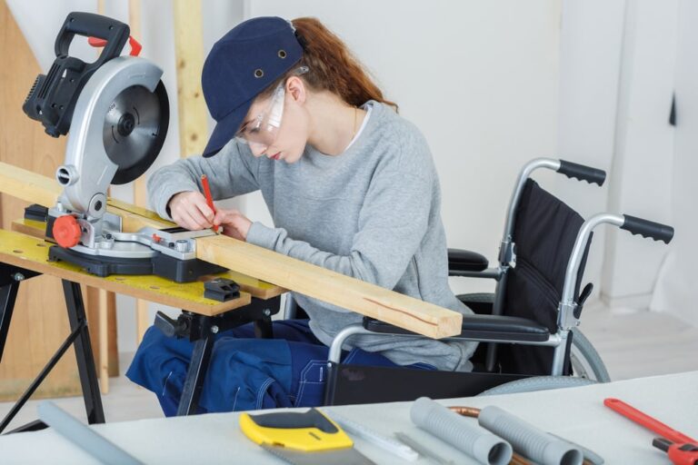 Person in a wheelchair working on a piece of wood with a circular saw on a workbench, wearing safety goggles and a cap.
