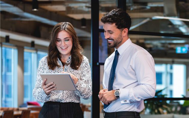 Two people in business attire stand in an office, engaged in conversation. One is holding a tablet.