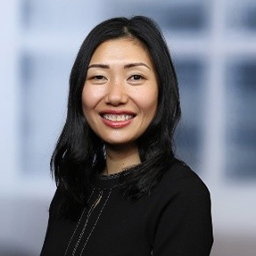 A woman with long black hair and a black top smiles at the camera. She stands in front of a blurred background.