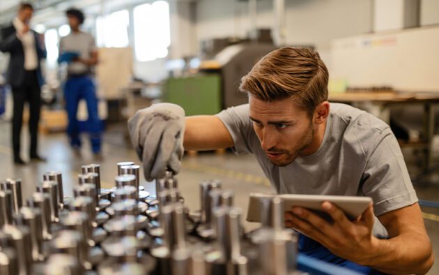 A man in a workshop inspects metal parts on a table, holding a tablet. Another person stands in the background.