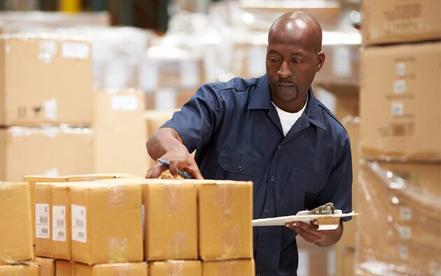 A warehouse worker in a navy shirt checks and inspects stacked cardboard boxes, holding a clipboard.