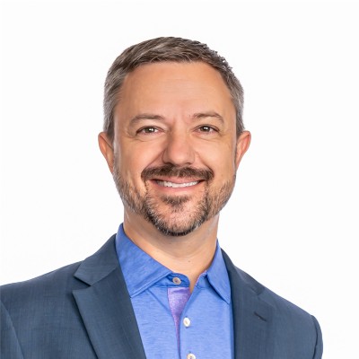 A man with short hair and a beard, wearing a blue shirt and a blue blazer, smiling at the camera against a plain white background.