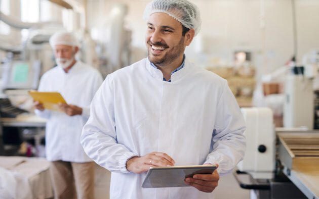 A man in a white coat and hairnet smiles while holding a tablet in a food processing facility. Another person in similar attire holds a folder in the background.