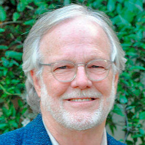 Elderly man with glasses and a beard smiles in front of lush green foliage.