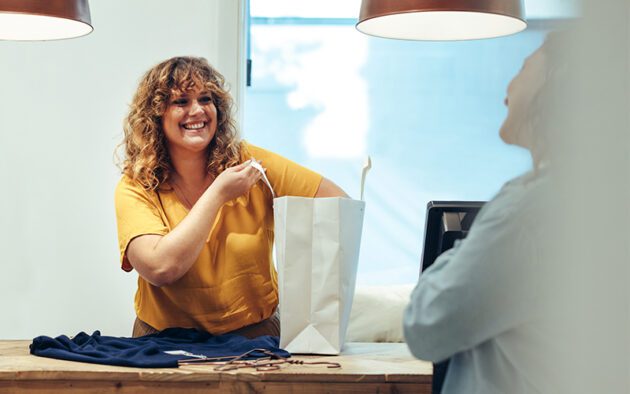 Smiling woman in a yellow top at a counter places an item into a white shopping bag.