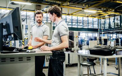 Two men in a factory setting discuss work at a desk with computers. Machinery and industrial equipment are visible in the background.