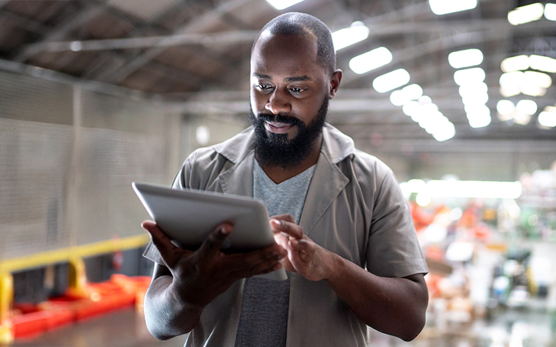 A man standing in a warehouse uses a tablet, focusing on the screen with shelves and industrial equipment blurred in the background.