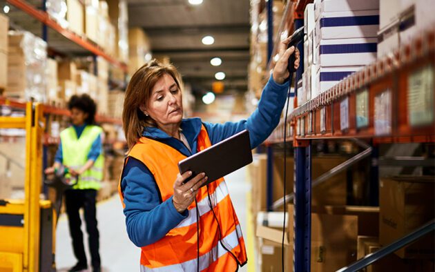 Woman in an orange vest scans items on a shelf in a warehouse, holding a tablet. Another person in a yellow vest is seen in the background.