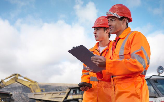 Two construction workers in orange safety gear and helmets stand outdoors, holding a clipboard, with machinery and a cloudy sky in the background.