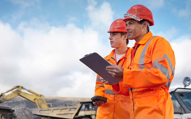Two construction workers in orange safety gear and helmets stand outdoors, holding a clipboard, with machinery and a cloudy sky in the background.