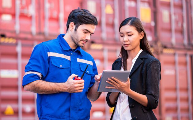 A male worker in blue overalls and a female colleague in business attire discuss something on a tablet in front of red shipping containers.