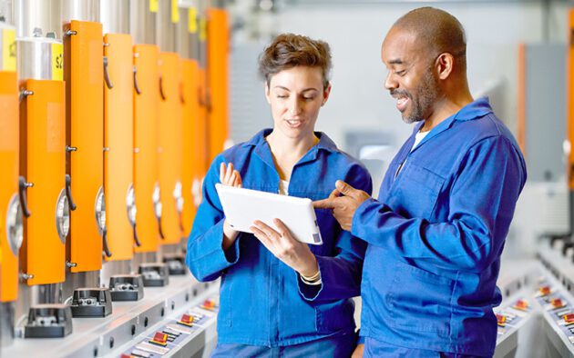 Two factory workers in blue uniforms discuss something on a tablet in an industrial setting with machines in the background.