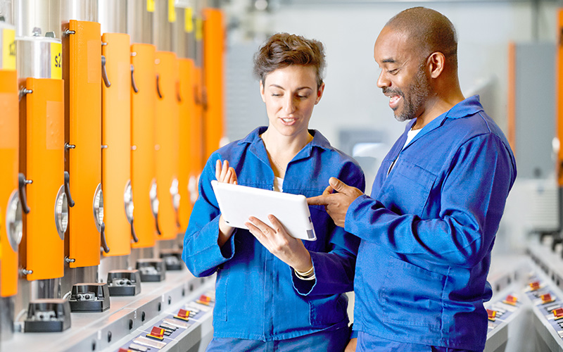 Two factory workers in blue uniforms discuss something on a tablet in an industrial setting with machines in the background.