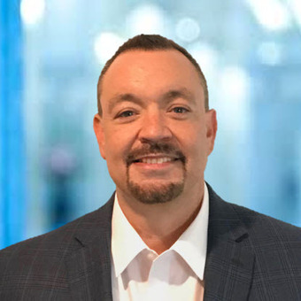 A man with a goatee, wearing a white shirt and a dark checkered blazer, stands in front of a blurred indoor background.