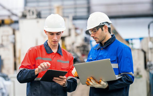 Two factory workers wearing helmets and protective clothing review information on a tablet and a laptop in an industrial setting.