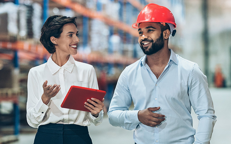 Two people, one holding a tablet and the other wearing a red hard hat, are talking and smiling in a warehouse setting.