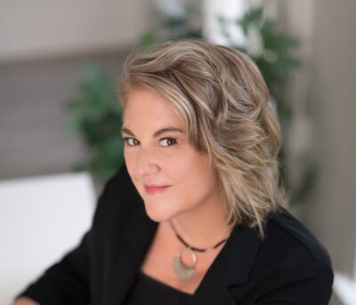 A person with short, wavy blonde hair wearing a black blazer and necklace, smiles slightly while sitting indoors.