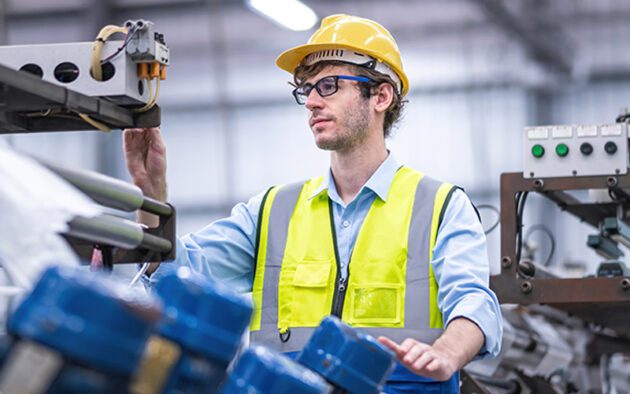 A worker wearing a yellow hard hat and neon safety vest operates machinery in an industrial setting.