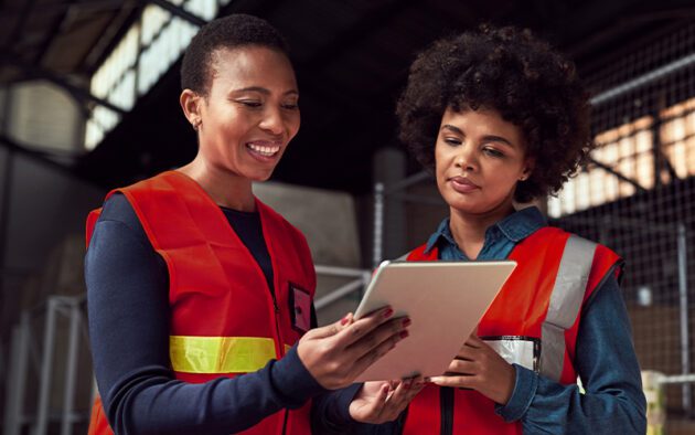 Two women wearing red safety vests stand indoors, looking at a tablet together. One smiles while holding the tablet, the other observes attentively.