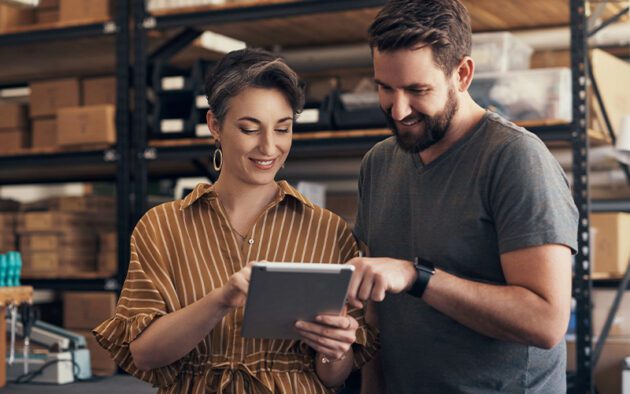 Two people stand in a storage room, smiling while looking at a tablet.