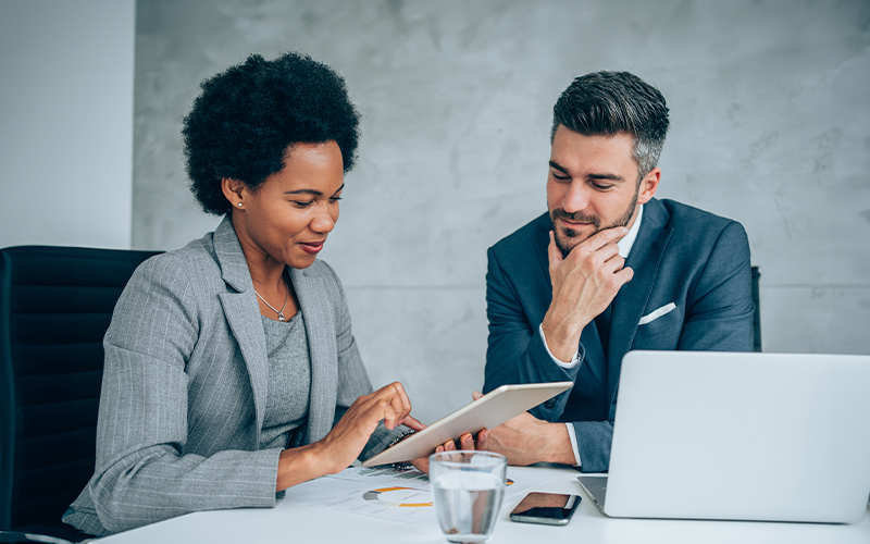 Two professionals in business attire are discussing something on a tablet at a desk, with a laptop and papers nearby.