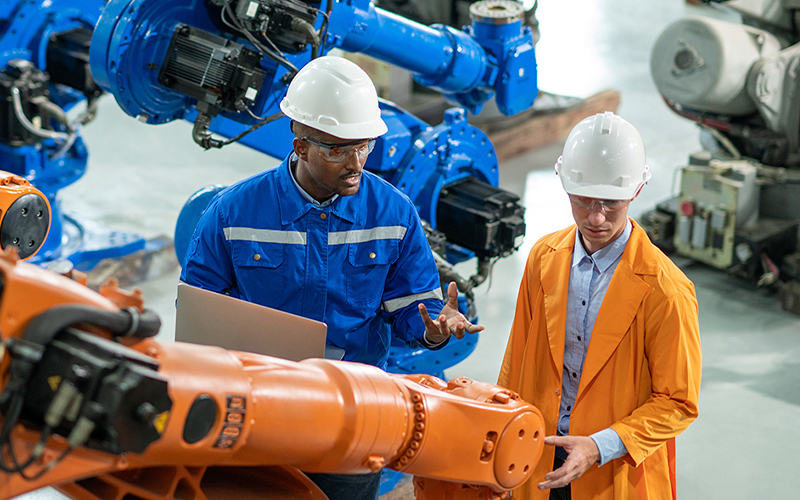 Two engineers in hard hats and safety gear inspect a robotic arm in an industrial setting, with one holding a laptop and the other wearing an orange coat.