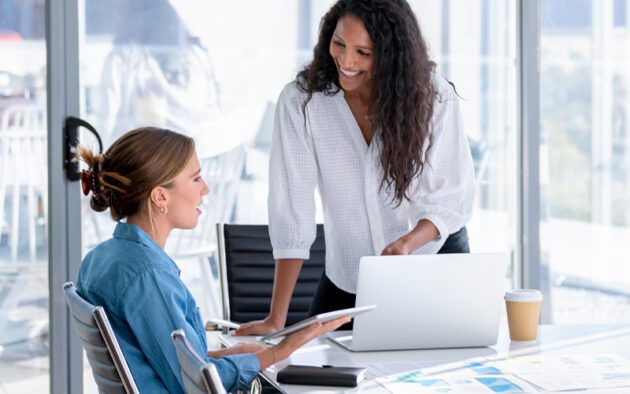 Two women in a meeting room, one sitting with a tablet and the other standing, smiling and engaged in conversation. A laptop and coffee cup are on the table nearby.