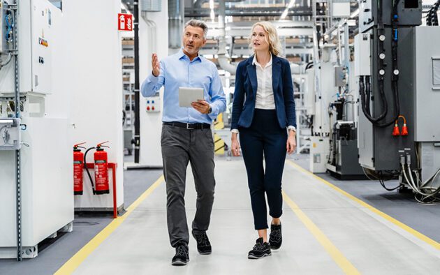 Two people in business attire walk and talk in a modern industrial facility, surrounded by machinery and fire extinguishers. One holds a tablet.