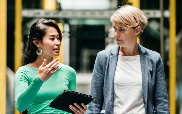 Two people in conversation, one holding a tablet. They appear to be in a professional or industrial setting.