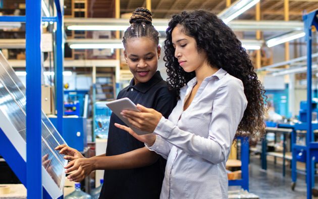 Two women in a factory setting, one holding a tablet and the other touching a glass panel, engaged in discussion.