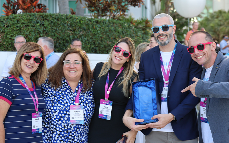 Five people in casual attire wearing name tags and sunglasses, one holding a blue bag, standing outdoors with greenery in the background.