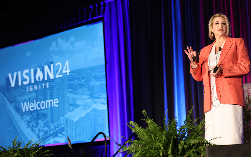A speaker in a pink blazer gestures on stage next to a large screen displaying "VISION24 IGNITE Welcome" against a blue background. Stage plants are visible in the foreground.