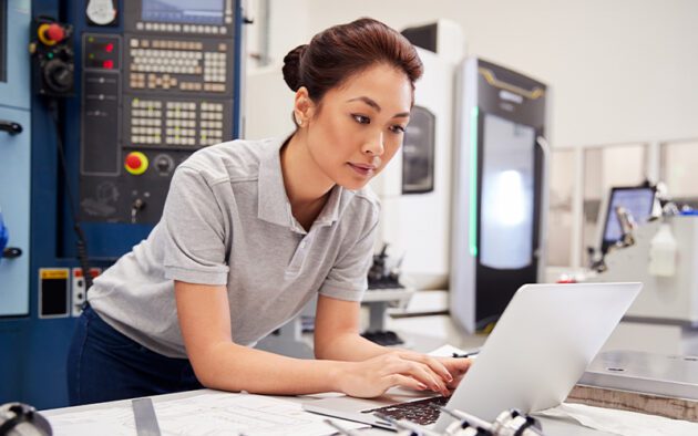 A woman wearing a grey polo shirt is working on a laptop in a laboratory or industrial setting with various machines in the background.