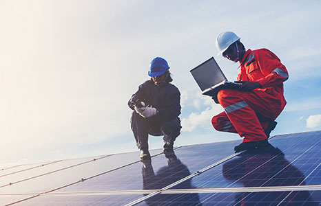 Two people wearing hard hats, one is working on a laptop while wearing reflective gear, kneeling on solar panels