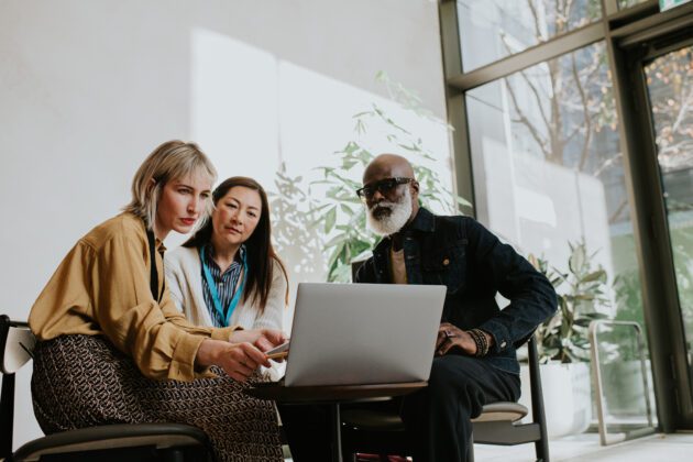 Three people sit around a laptop. One woman is presenting to the group.