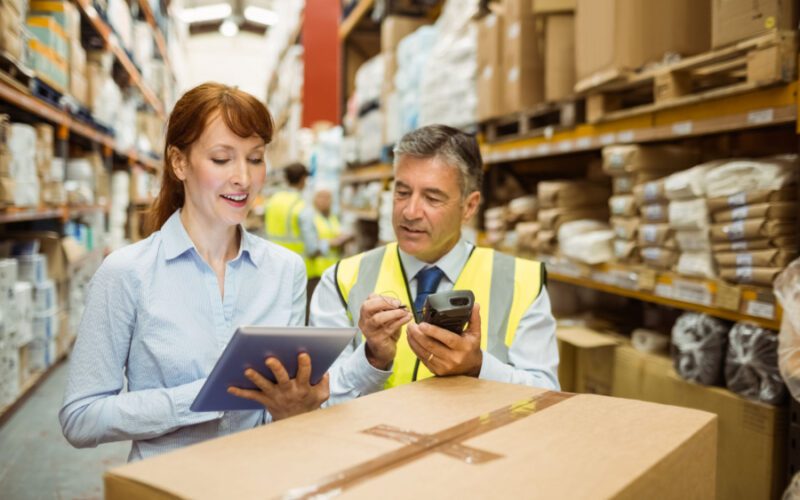 Two warehouse employees in safety vests check a tablet and scan a package in an aisle lined with shelves of inventory.