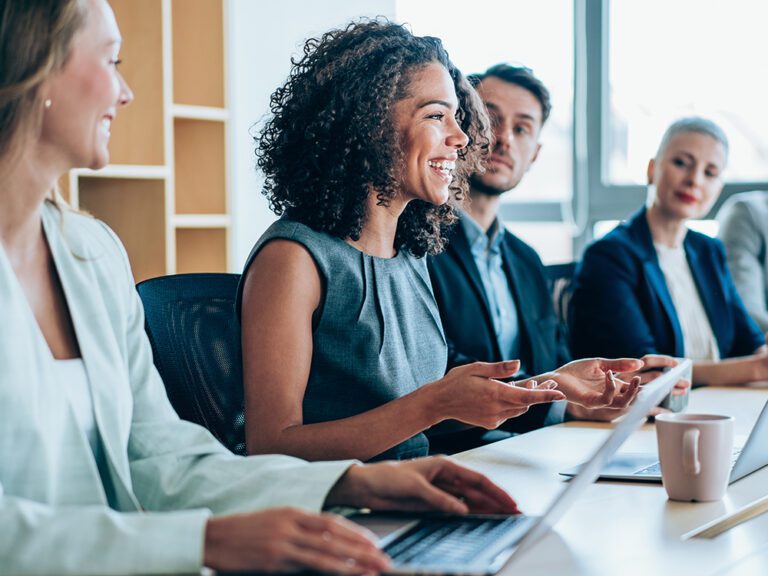 A diverse group of professionals sit around a conference table. A woman in the center is speaking, while others listen attentively. Laptops and coffee cups are on the table.