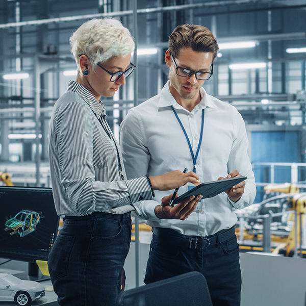 Two people in an industrial setting review documents on a tablet. They stand next to a computer displaying a vehicle design.