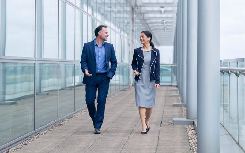 Two business professionals, a man and a woman, walk and talk in a glass-walled walkway. Both are dressed in business attire, with the man in a blue suit and the woman in a gray dress with a navy blazer.