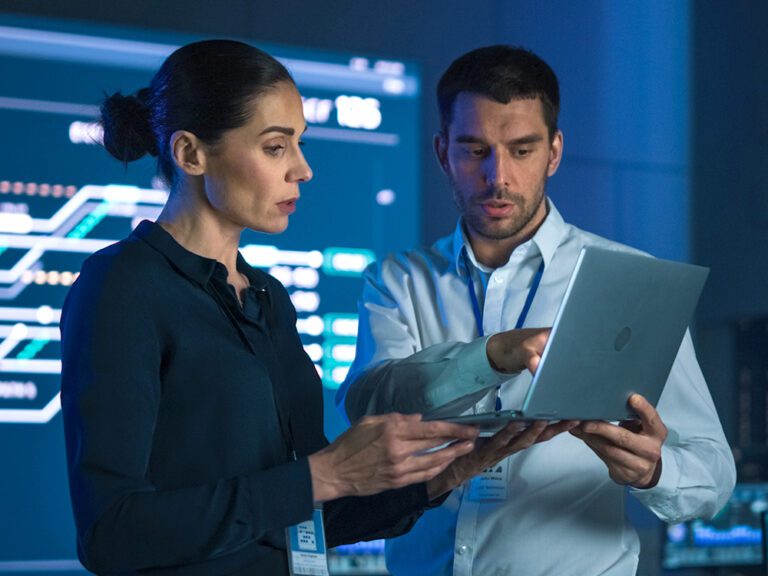 Two individuals are examining a laptop screen while standing in front of large monitors displaying data in a technological setting.