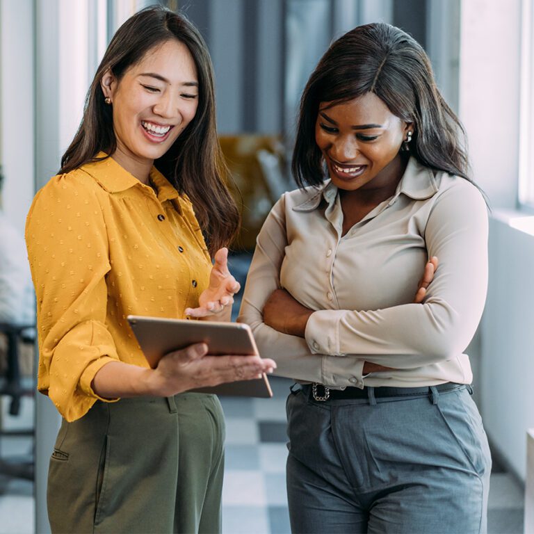 Two women stand in an office, looking at a tablet together while smiling. One wears a yellow blouse, and the other is dressed in a light grey blouse.