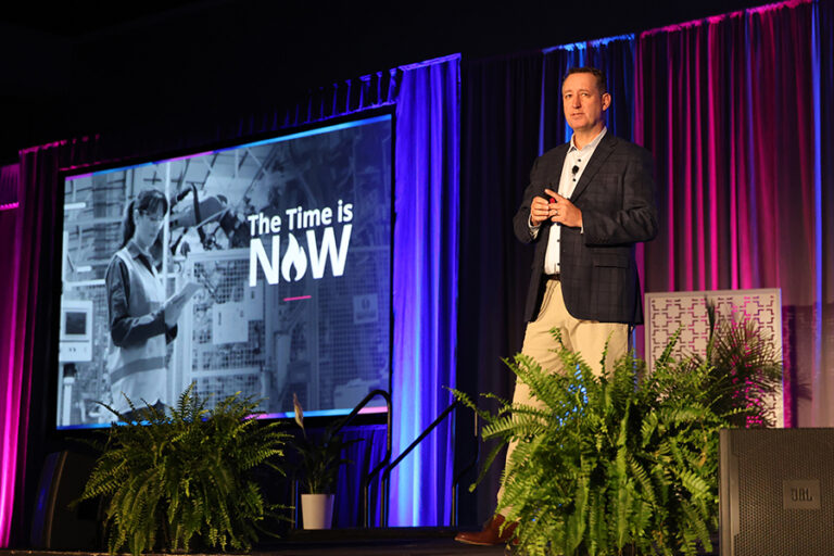 A man in a suit speaks on stage in front of a screen with the text "The Time is NOW," featuring an industrial worker. The stage is decorated with plants and purple lighting.