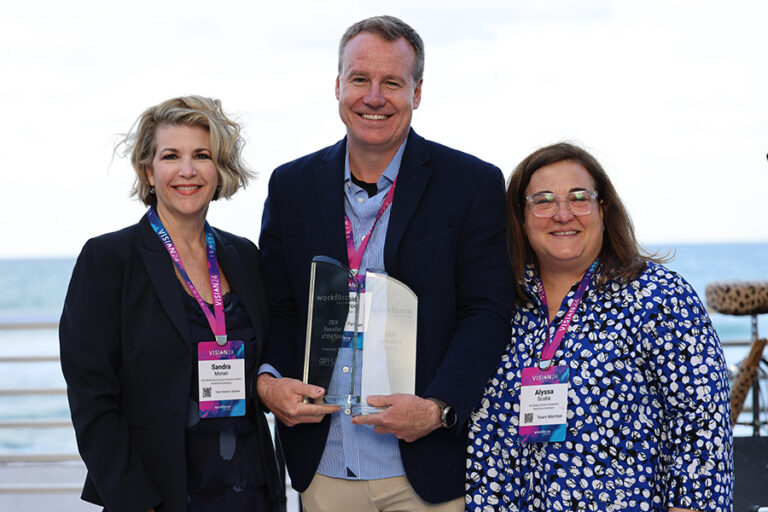 Three people stand together outdoors, each wearing name tags and holding an award. They are smiling at the camera with a background of the sea and sky.