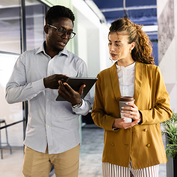 Two colleagues, one holding a tablet and the other a coffee cup, discuss work while walking in a modern office space.