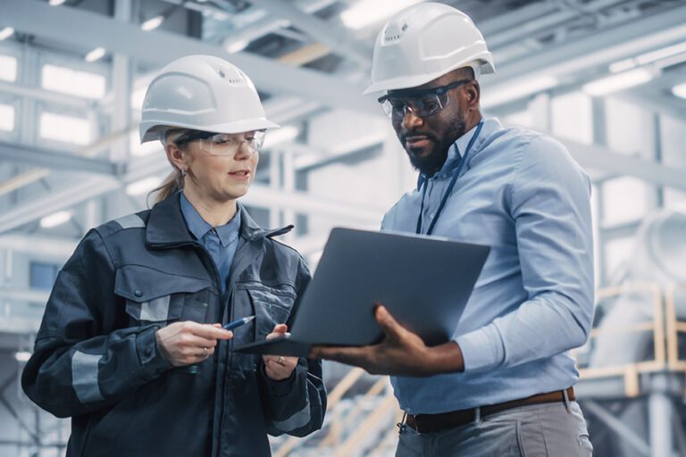 Two engineers wearing safety helmets and glasses review information on a laptop in an industrial setting. One is holding a pen and pointing at the screen.