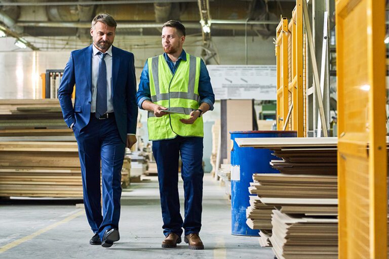 Two men walking in a warehouse. One is wearing a blue suit and holding a folder, the other is in a neon safety vest and holding a tablet. They appear to be discussing something related to the workspace.