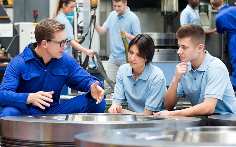 A person in a blue jumpsuit explains something to two young individuals seated beside machinery in a workshop. Other individuals are working in the background.