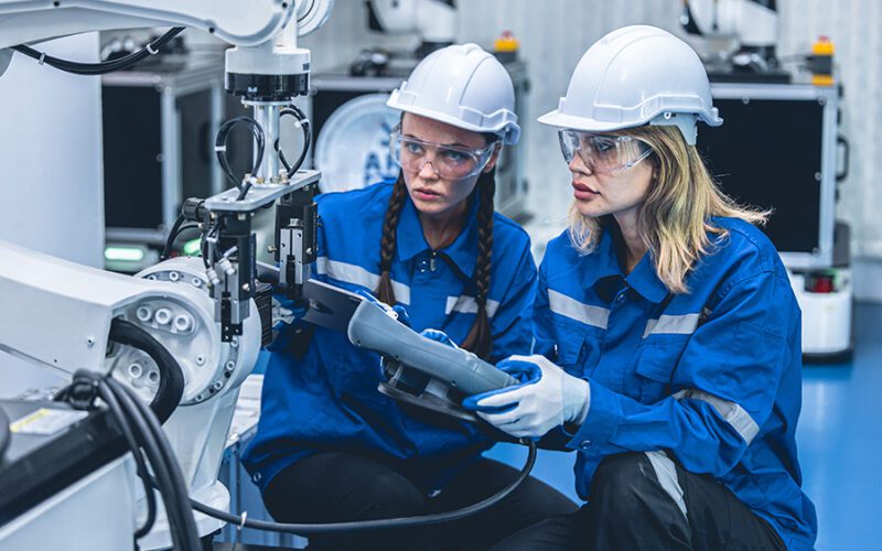 Two workers wearing blue uniforms, safety helmets, and goggles are operating a robotic arm in a factory setting. One holds a control device as they both focus on the machinery.