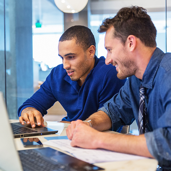 Two men are seated at a desk, working together on a laptop, with documents and another laptop in front of them in a modern office setting.